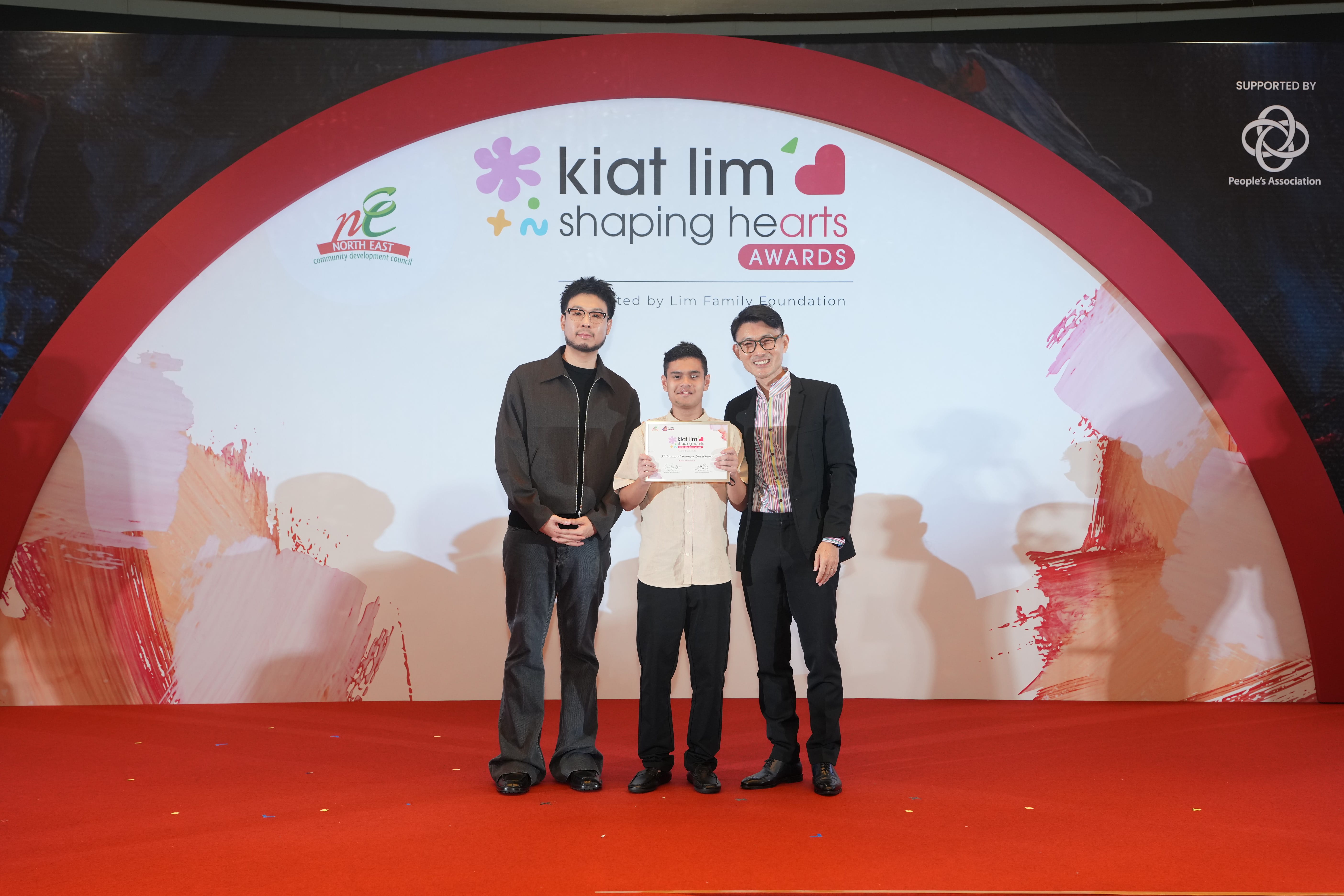 Award recipient standing on stage with the presenter, holding their certificate and posing for a group photo during the award ceremony, with a backdrop reading ‘Kiat Lim-Shaping Hearts Awards'.